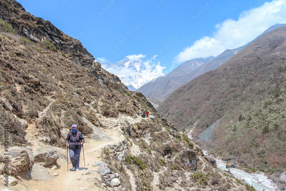 Fototapeta premium Middle-aged woman walks on footpath from Everest base campin Himalayas. Everest and Lhotse mountain peaks are visible in the background. Imja Khola river flows in valley. Trekking in Nepal theme.