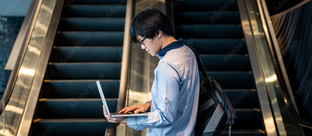 Asian man with glasses and bag holding laptop computer standing in front of escalator in public building. Working anywhere concept