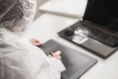 Girl in medical mask and white transparent protective suit sits in quarantine at home and works at computer and graphics tablet. Designer, artist, architect at remote work in a pandemic covid.