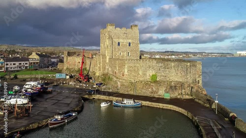 Medieval Norman Castle and harbor in Carrickfergus near Belfast in sunset light. Aerial 4K flyby video with  marina, breakwater and town. Blue sky and dark stormy clouds