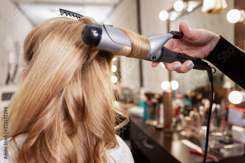 Blonde girl in a beauty salon doing a hairstyle. Twisting locks of hair on a styler. Close-up.