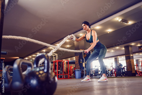 Caucasian fit woman dressed in sportsoutfit posing with battle ropes at gym. © zamuruev