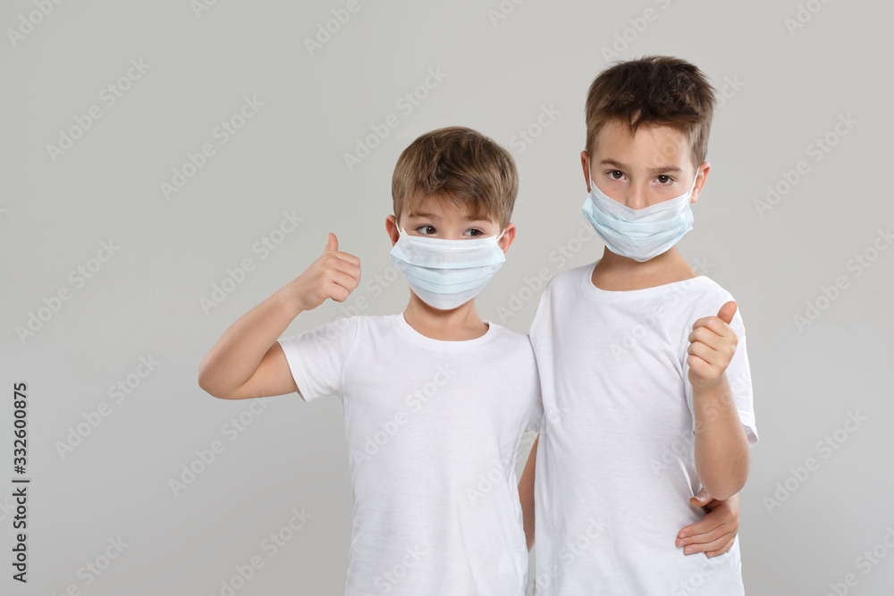 Two cute little boys in white clothes and protective medical masks show thumb up, isolated on a gray background.