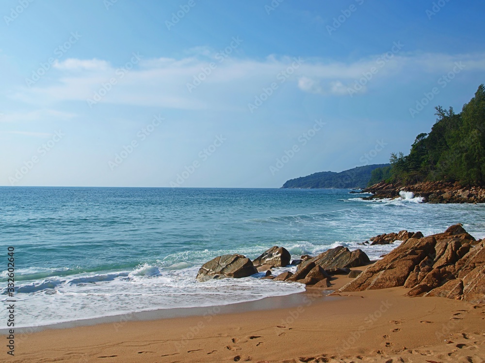 Sea coast, waves, rocks and stones on a sandy wild beach. Sea bay, coastline and hill of island on the horizon. Blue sky with white clouds.