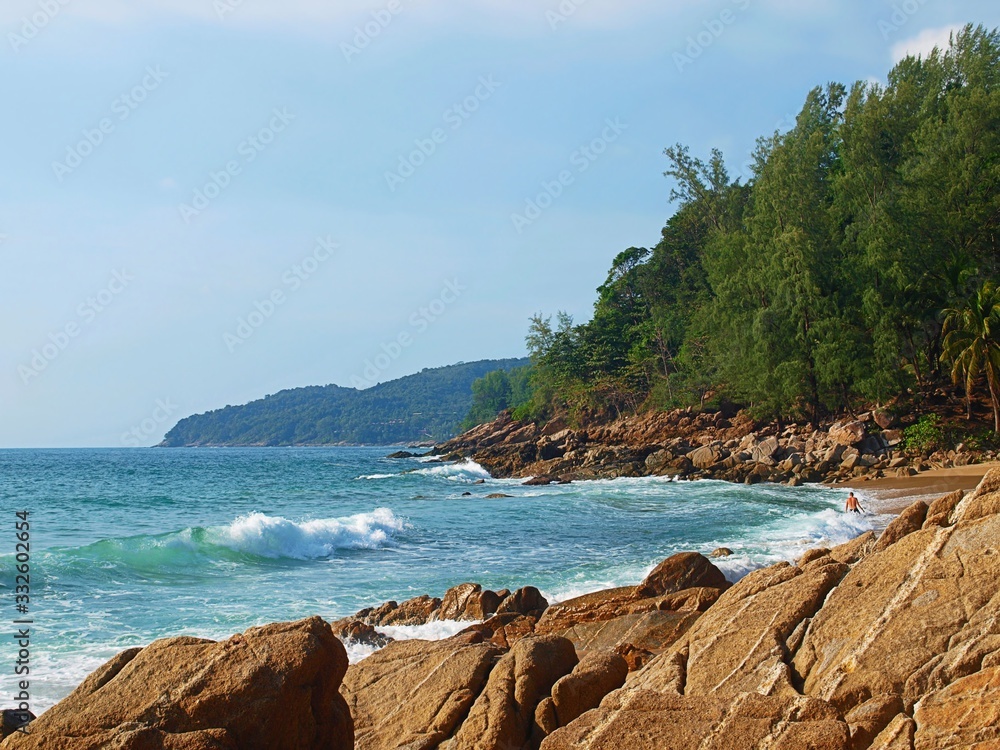 Fototapeta premium Panoramic view of rocky and sandy shore of a wild beach. Landscape of sea bay, the azure water and the crest of a wave rolling to the coast. Hills covered with green forest at the horizon.