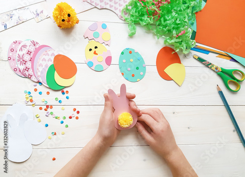 The child makes crafts with his own hands for the Easter. Colorful handmade from multi-colored paper. Scissors, cardboard, eggs, chicken, rabbit. Art creativity on a wooden table. Top view, copy space