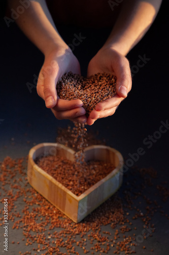 Children's hands pour buckwheat into a heart-shaped wooden plate. Buckwheat is poured into a storage tank. Buckwheat in the hands close-up.