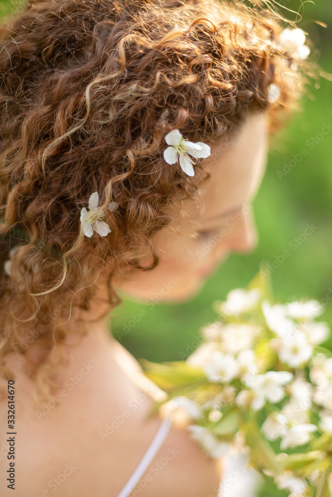 Naklejka premium Back view. A gorgeous tender girl with white flowers in curly hair holds a flowering branch in her hand. Inspiration of spring and summer. Perfume, cosmetics concept.