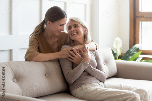 Loving adult daughter hugging older mother, standing behind couch at home, family enjoying tender moment together, young woman and mature mum or grandmother looking at each other, two generations