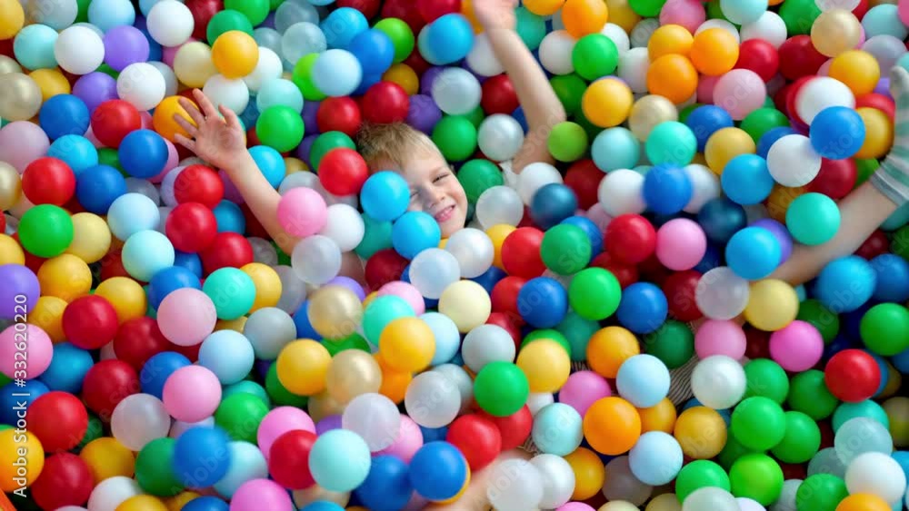Blonde little boy scatters balls in different sides, lying on multi coloured plastic balls in big dry paddling pool in playing centre. Smiling at camera. Having fun in playroom. Leisure Activity. Blonde little boy scatters balls in different sides, lying on multi coloured plastic balls in big dry paddling pool in playing centre. Smiling at camera. Having fun in playroom. Leisure Activity.