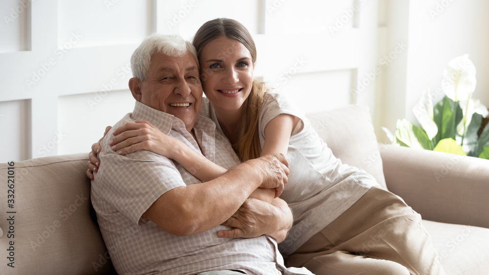 Portrait happy older father and adult daughter hugging, looking at camera, posing for photo, beautiful young woman with smiling mature dad or grandfather sitting on cozy sofa at home, two generations