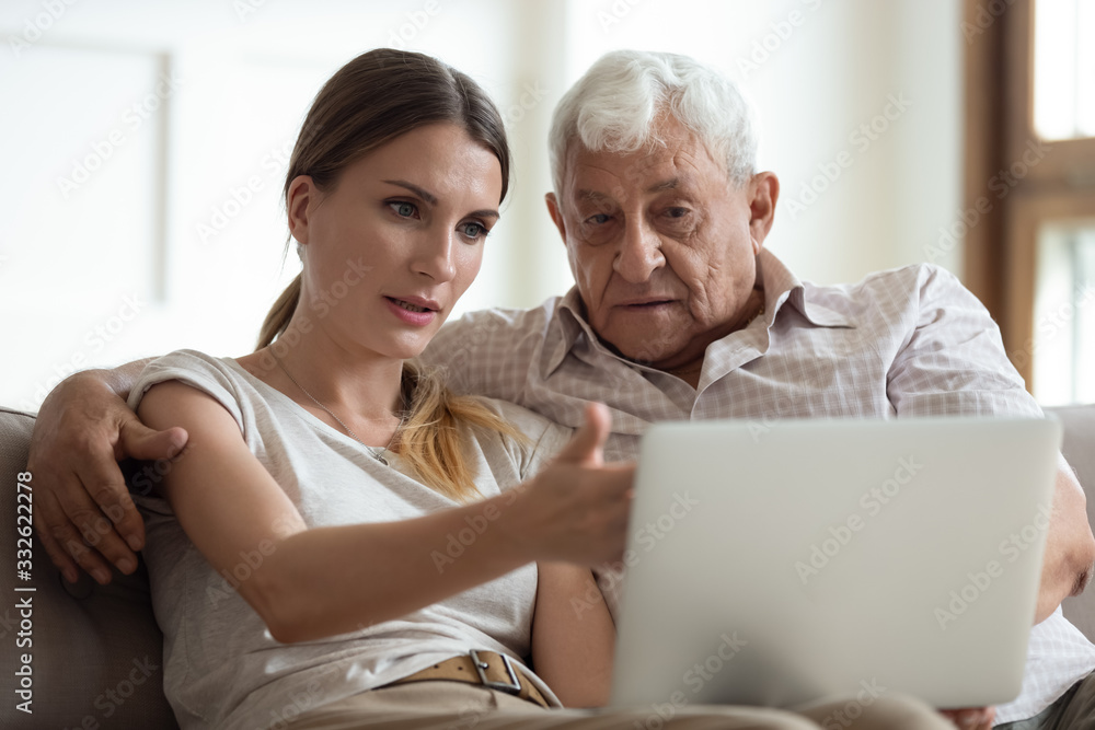 Serious adult daughter helping older father with laptop close up, pointing at screen, explaining, focused young woman teaching mature dad or grandfather to use computer, sitting on couch together