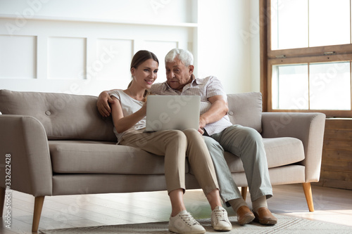 Smiling adult daughter teaching older father to use computer, pointing at screen, explaining, happy young woman and mature dad or grandfather hugging, using laptop together, sitting on couch at home