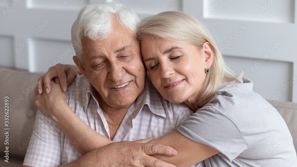 Close up older husband and wife hugging with closed eyes, senior family enjoying tender moment, adult middle-aged daughter embracing mature father, expressing love and gratitude, two generations
