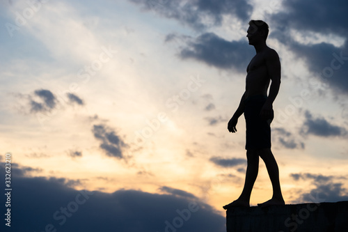 Fototapeta Naklejka Na Ścianę i Meble -  Silhouette of man against background of clouds and sunset. He stands on edge of roof. He is ready to take a step. Concept of Risk and courage