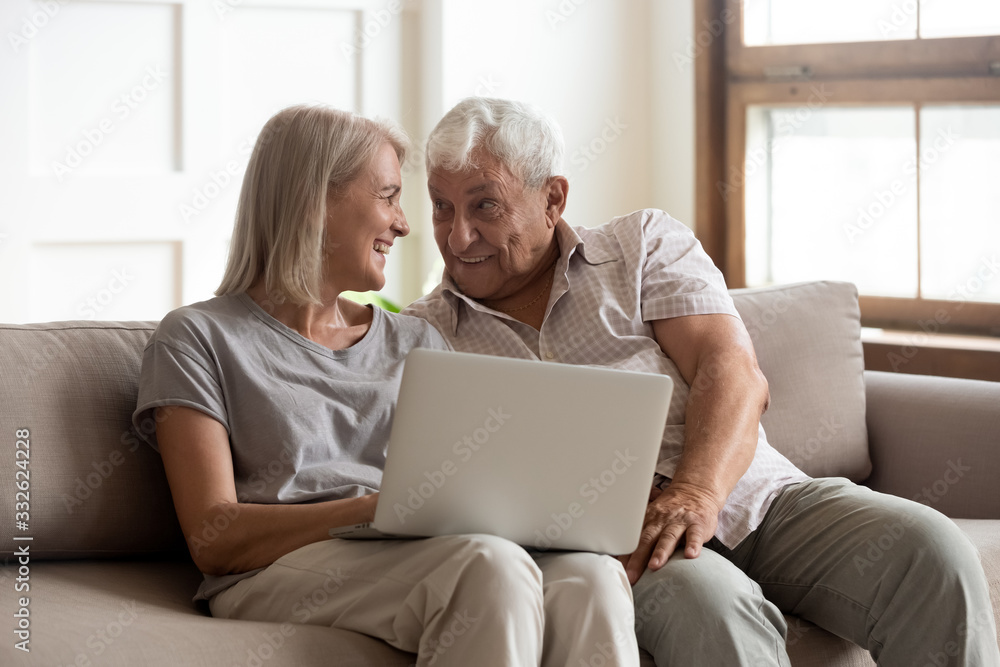 Happy older wife and husband using laptop together, having fun, smiling mature grandparents shopping or chatting online, sitting on cozy sofa at home, looking at each other, enjoying weekend Happy older wife and husband using laptop together, having fun, smiling mature grandparents shopping or chatting online, sitting on cozy sofa at home, looking at each other, enjoying weekend