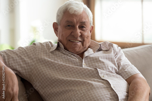 Head shot portrait close up smiling older retired man sitting on couch at home, happy friendly grandfather looking at camera, mature grey haired male posing for photo on sofa, feeling positive
