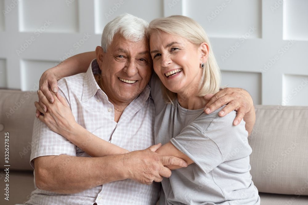 Head shot portrait older wife and husband with healthy toothy smiles hugging, looking at camera, sitting on cozy sofa at home, happy adult middle-aged daughter embracing mature father, family photo