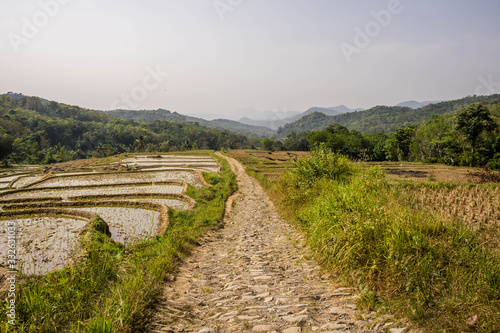 West Java countryside stone hand paved road, rice paddies and hills in haze in distance. Human hand made stone path solution in flood season