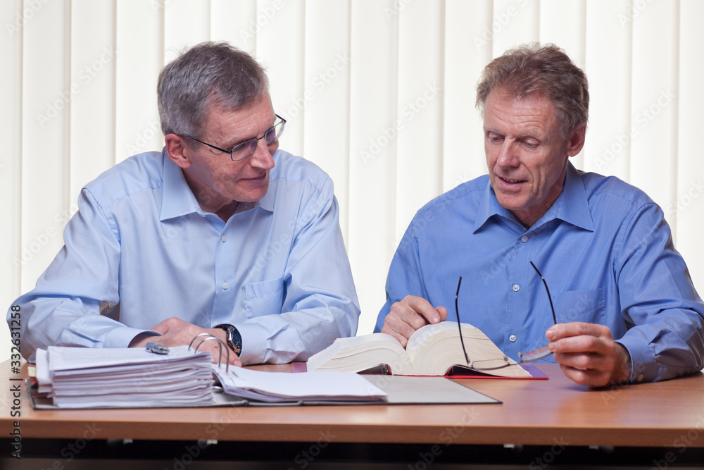 Two mature businessmen or partners readin in a book sitting at a desk with folder and book Two mature businessmen or partners readin in a book sitting at a desk with folder and book