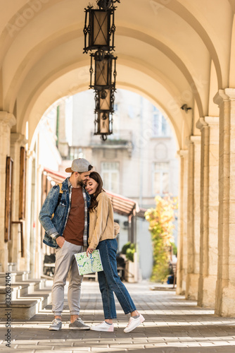 Selective focus of boyfriend with hand in pocket hugging girlfriend with map in city