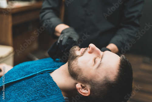 Barbershop. The client in the master’s chair in the barbershop, the barber applies oil and cosmetics to the client’s beard. Male beauty shop. Healthy lifestyle and beauty.
