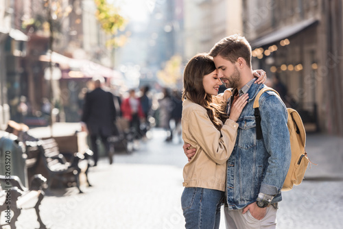 Boyfriend with hand in pocket and girlfriend hugging with closed eyes in city in Europe