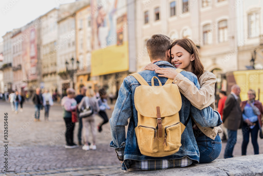Boyfriend with backpack and girlfriend with closed eyes hugging and smiling in city