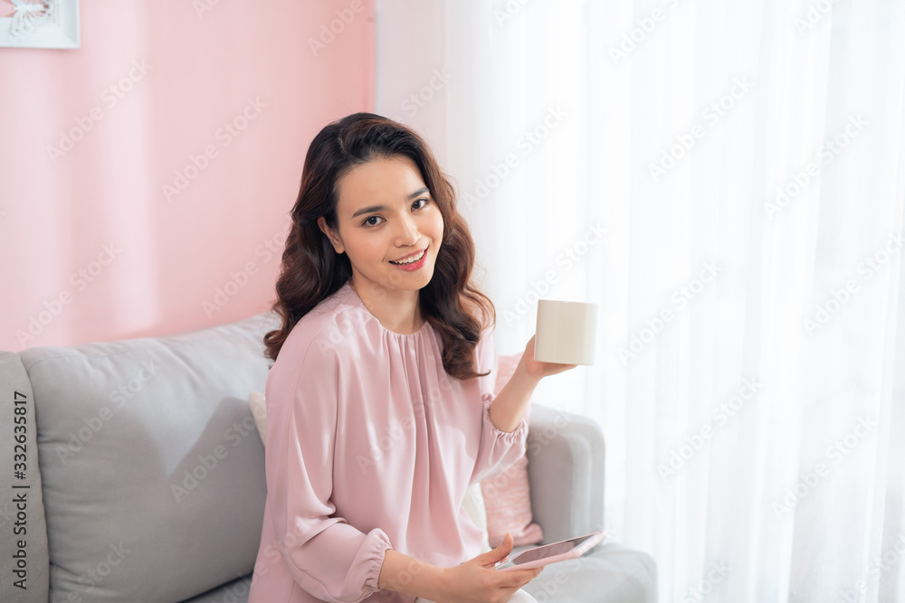 Happy young Asian girl using phone and drinking coffee, tea on sofa. Happy young Asian girl using phone and drinking coffee, tea on sofa.