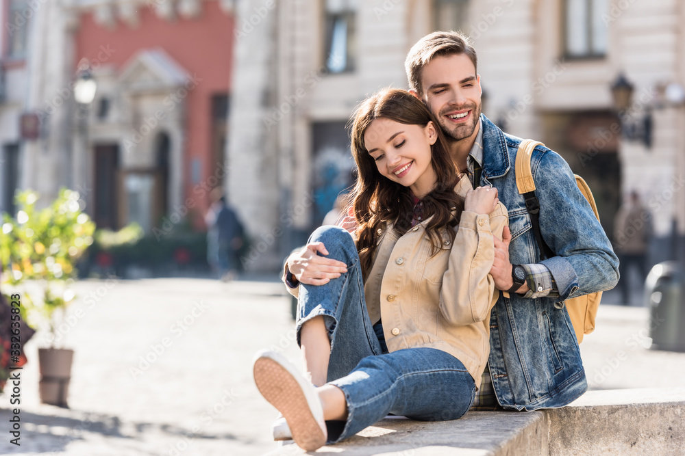 Selective focus of boyfriend hugging girl sitting on stone surface and smiling in city