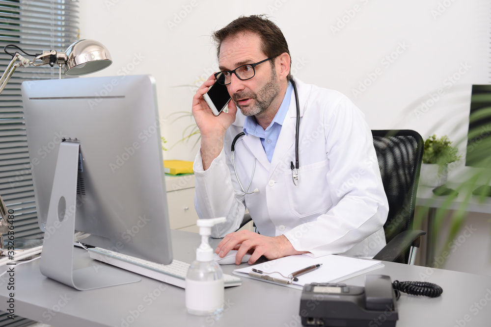 mature male doctor in medical office at desk on computer with stethoscope struggling against covid19 and coronavirus