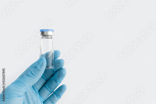 Doctors hand in blue rubber glove holding medicine bottle with blue cover and clear fluid on grey background. Coronavirus vaccine development. 2019-nCoV liquid drug, virus antidote. Copy space.
