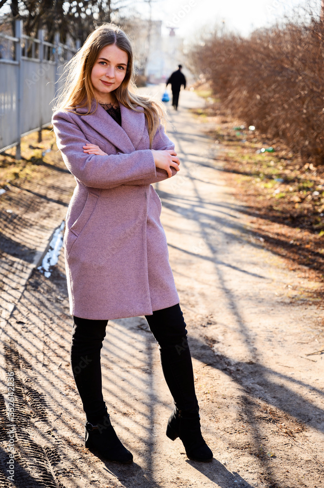 Full-length portrait of a pretty smiling posing blonde Caucasian girl with long beautiful hair outdoors in a pink coat in a park against a tree in the afternoon on a sunny day.