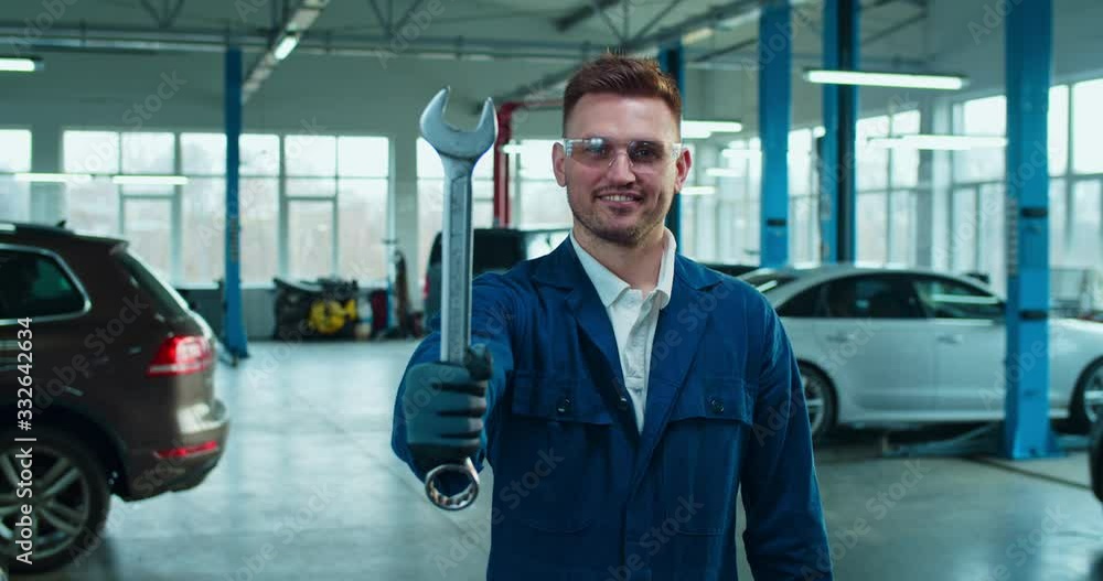Portrait of happy cheerful Caucasian handsome man auto mechanic smiling to camera and showing wrench in garage. Young guy worker in overalls and goggles holding tool at cars service. Portrait of happy cheerful Caucasian handsome man auto mechanic smiling to camera and showing wrench in garage. Young guy worker in overalls and goggles holding tool at cars service.