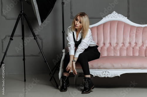Beautiful girl in a white shirt and black overalls on a pink sofa in a photostudio on fashion photography