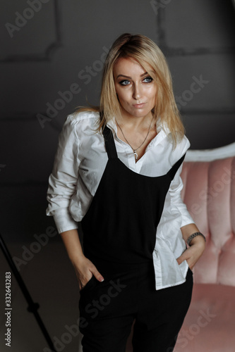 Beautiful girl in a white shirt and black overalls on a pink sofa in a photostudio on fashion photography