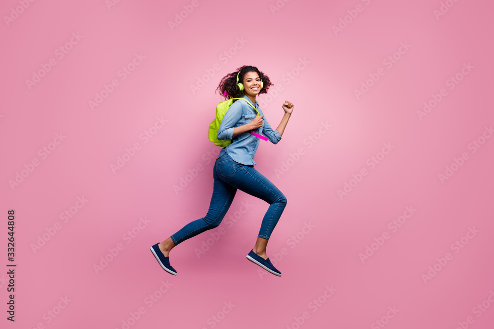Full length profile side photo of cheerful afro american girl college student jump run hurry lecture listen music headset wear denim jeans shirt bag travel isolated pink color background