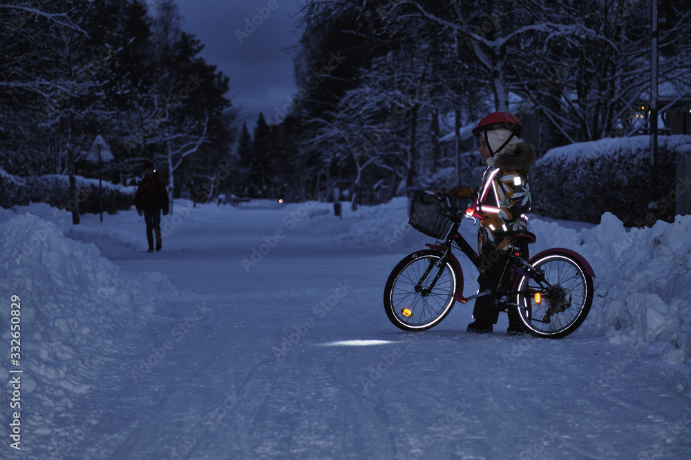 Girl on a bicycle in a winter evening. Light is reflected from clothing reflectors and bicycle wheels. Safe cycling in the dark time. - obrazy, fototapety, plakaty 