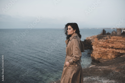 Young stylish woman 24-26 year old posing at sea shore at the top of rock outdoors. Looking forward. Spring season. 20s.