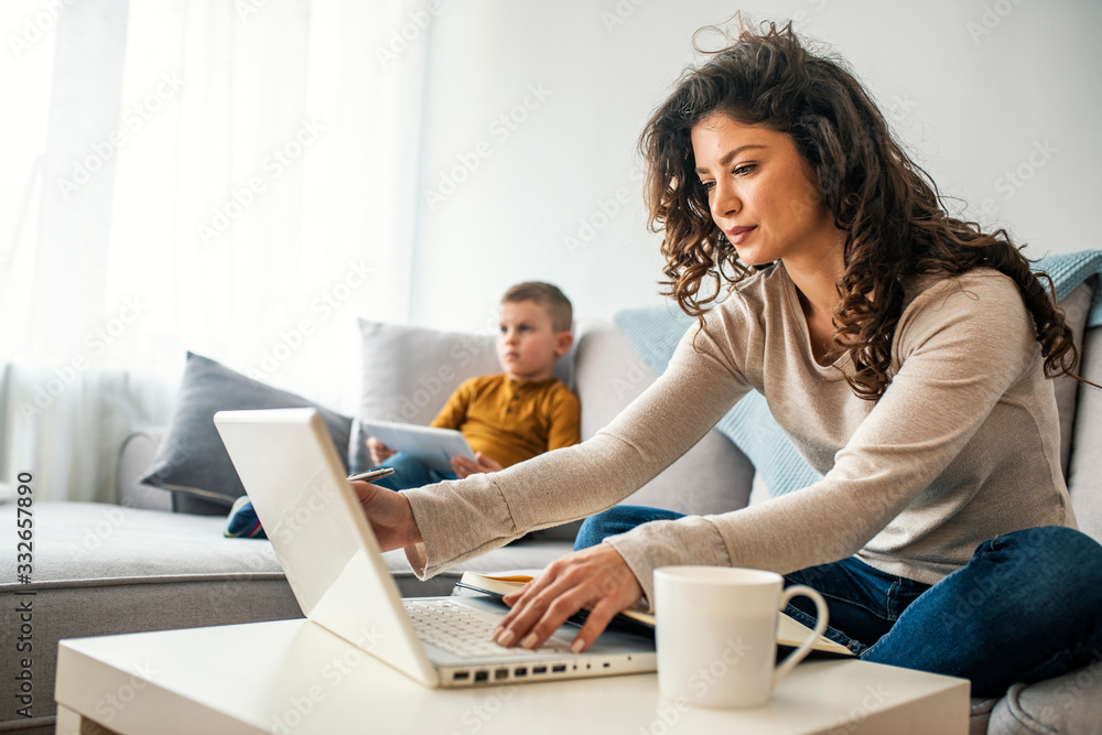 Smiling mom working at home with her child on the sofa while writing an email. Young woman working from home, while in quarantine isolation during the Covid-19 health crisis Smiling mom working at home with her child on the sofa while writing an email. Young woman working from home, while in quarantine isolation during the Covid-19 health crisis