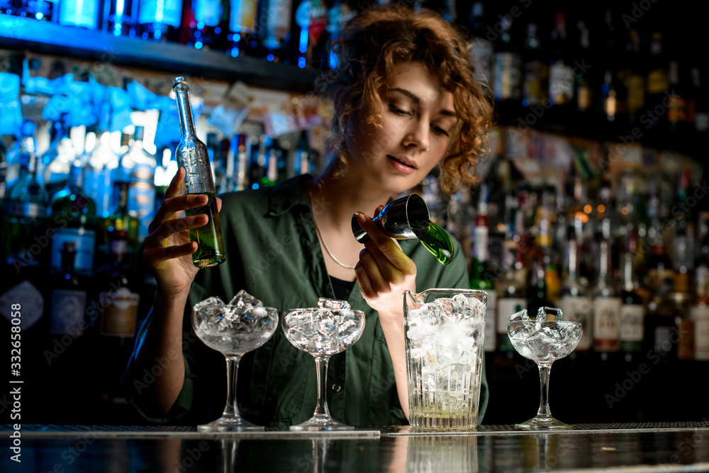 young woman bartender pours green drink into large glass used beaker.