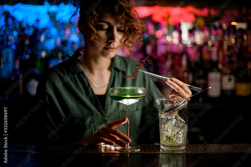 Young handsome woman bartender holds glass with cocktail and carefully decorates it