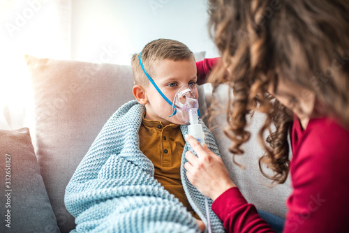 Woman with son doing inhalation with nebulizer at home. Causian little boy making inhalation with nebulizer. Child having respiratory illness helped by mother with inhaler photo