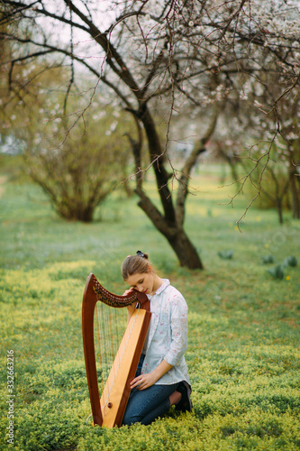 Valokuva Woman harpist sits on grass and plays harp among blooming apricot trees