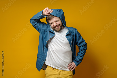 Attractive Caucasian bearded man in a raincoat puts on a hood and smiles cute. Isolated on a yellow background. © koldunova