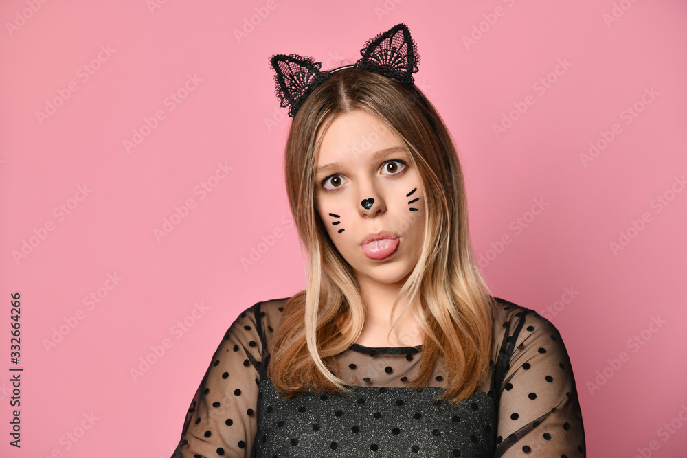Blonde teenager in black dress, headband like cat ears, face painting. She showing her tongue, posing on pink background. Close up