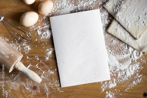 Baking ingredients. Bowl, eggs, flour, egg cattle, rolling pin and egg shell on a wooden board on top. A sheet of paper and ingredients for baking