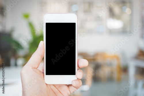 woman hand holding a smartphone with Mockup, a blank black screen in the white coffee shop. Close up
