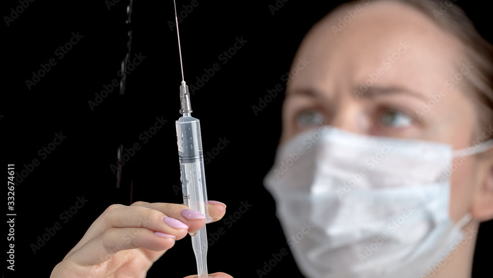 Female doctor in medical gown with sterile face mask looks to a drops of medicine vaccine that dripping with drops from syringe in her hands. Medicine and vaccination. Anti-virus human health concept. Female doctor in medical gown with sterile face mask looks to a drops of medicine vaccine that dripping with drops from syringe in her hands. Medicine and vaccination. Anti-virus human health concept.