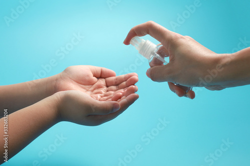 Teacher applying alcohol gel spray or antibacterial soap sanitizer on child's hands against blue background. 